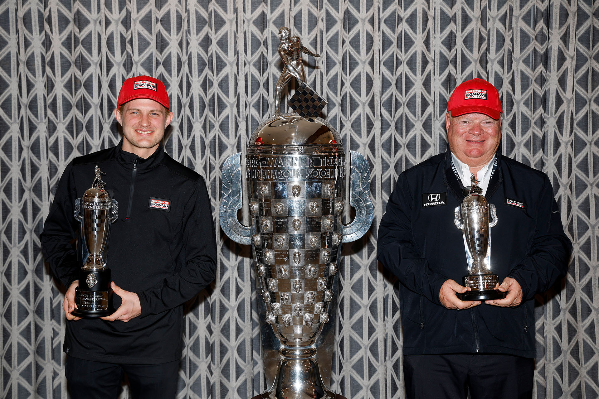 Two men in red hats stand next to large silver trophy while holding smaller trophies